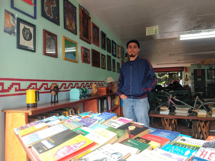 Mesa con libros, Ricardo Venegas de pie y cuadros que cuelgan en una pared. 